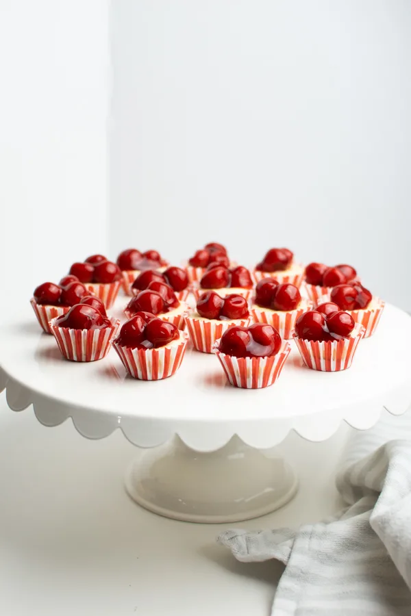 Several mini cheesecake bites topped with cherry pie filling on white cake plate.