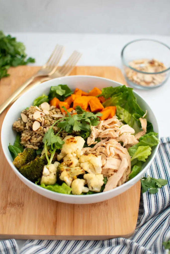 Chicken power bowl with roasted veggies and lettuce on wood cutting board with gold forks in background.