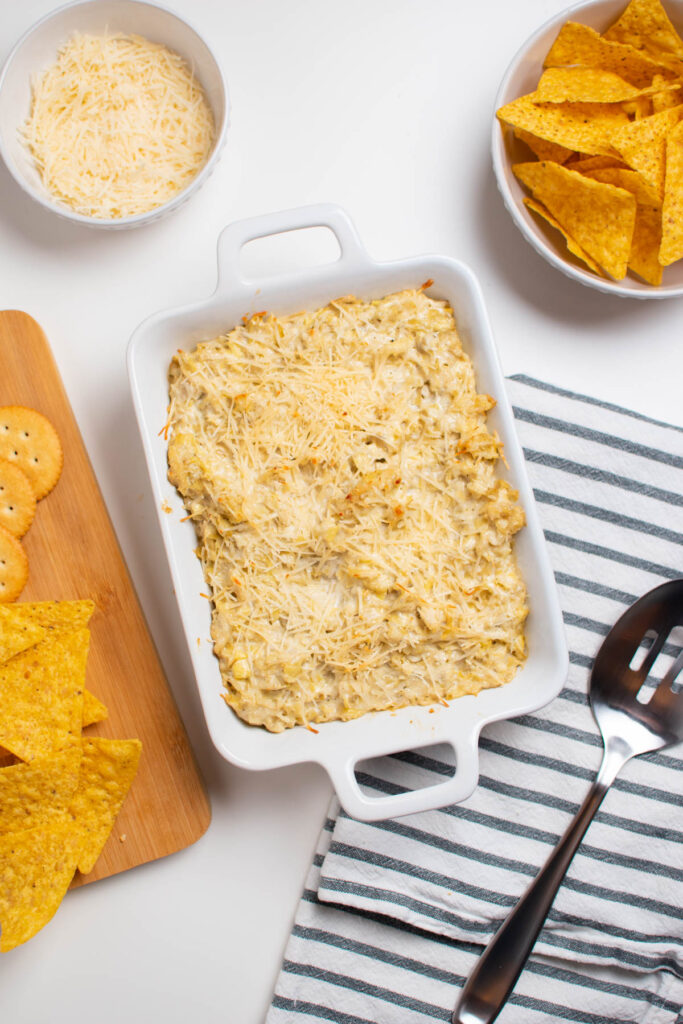 Hot artichoke dip in a small white baking dish with crackers and chips nearby.