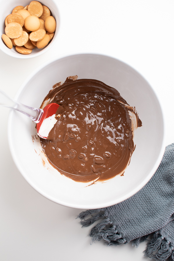 Melted chocolate in large white mixing bowl with red spatula next to bowl of wafers.
