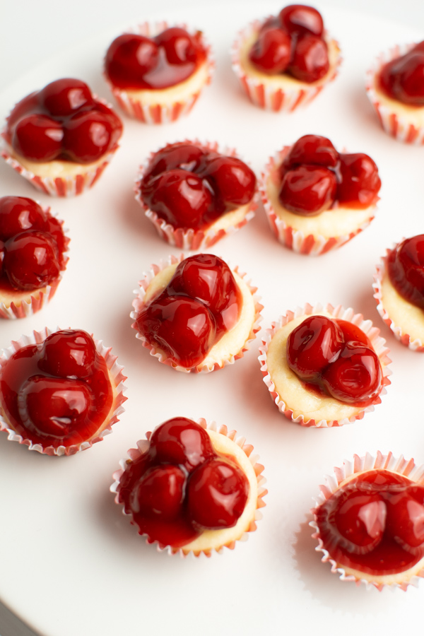 Mini cheesecakes in red and white cupcake liners topped with cherries on white plate.