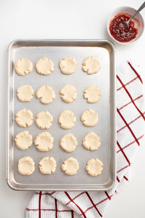 Thumbprint cookies on large metal baking sheet next to bowl of jam.