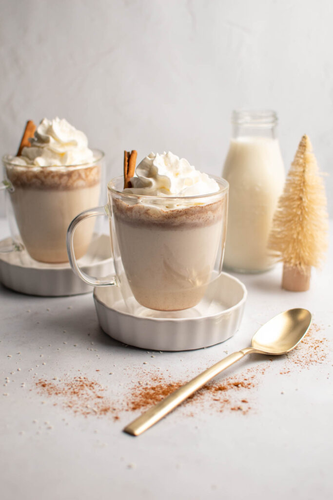 Two mugs of white hot chocolate on small dishes with gold spoon nearby.