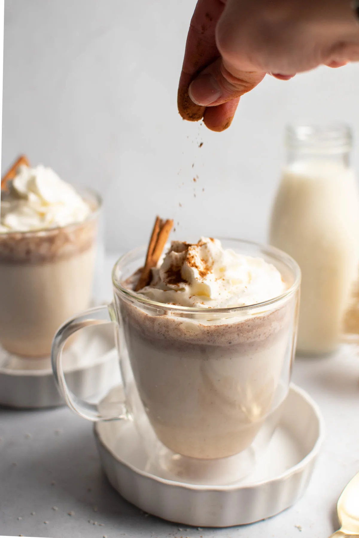Hand sprinkles cinnamon on mug of white chocolate hot cocoa with whipped cream and cinnamon stick.