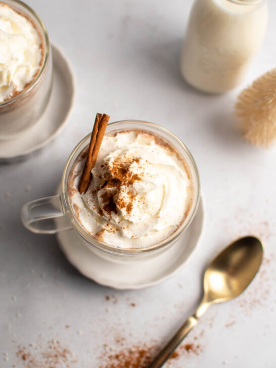 Mug with white hot chocolate, whipped cream, cinnamon, and cinnamon stick on table with spoon nearby.