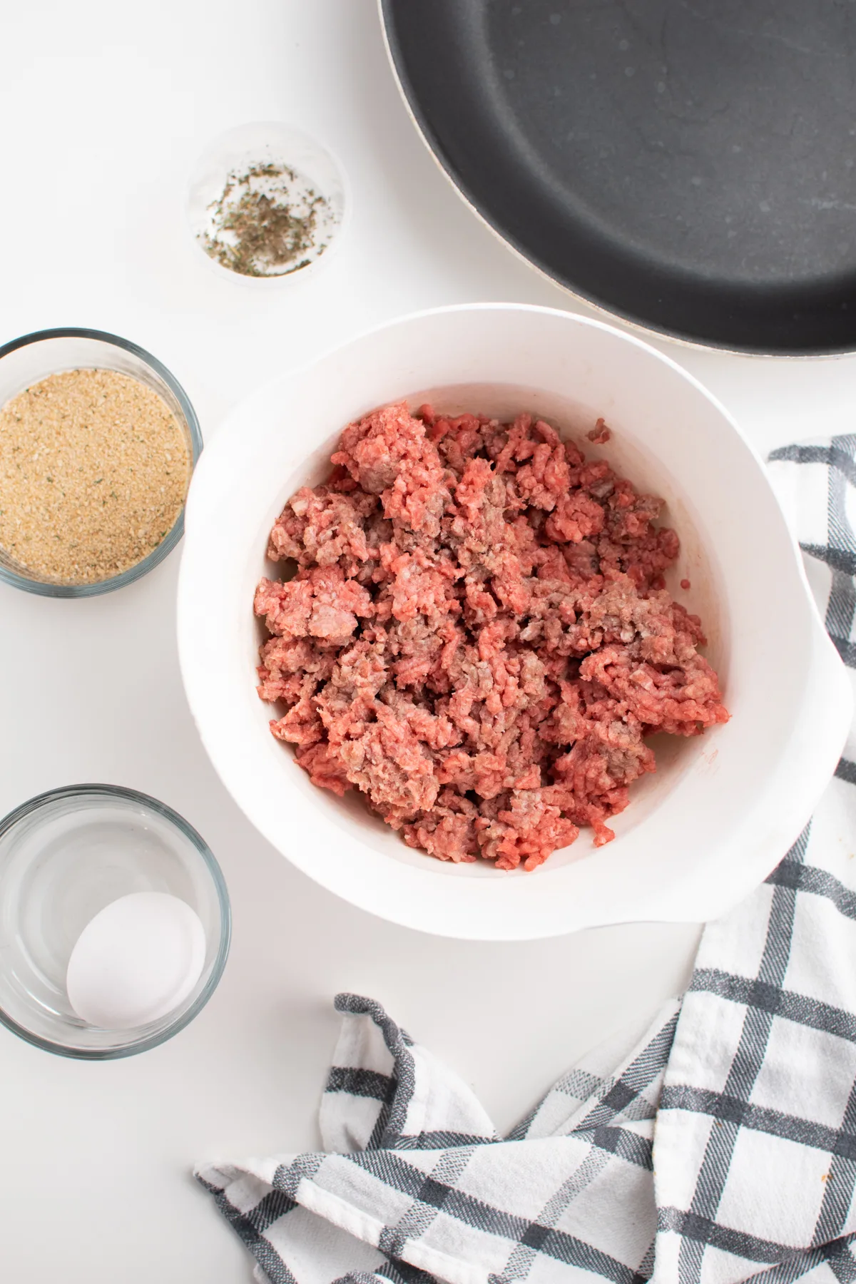 Ground beef in white mixing bowl next to egg, bread crumbs and seasonings.