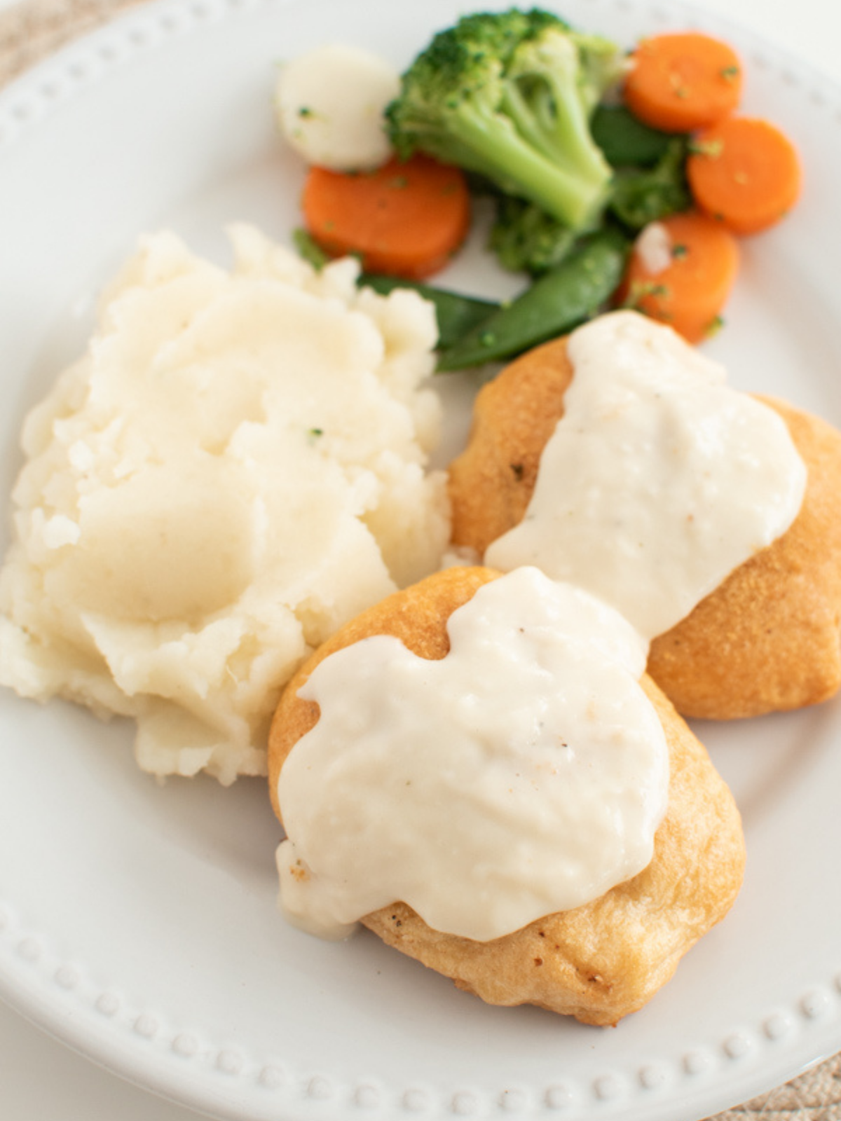 Chicken pillows with gravy on a white plate next to side of mixed vegetables.