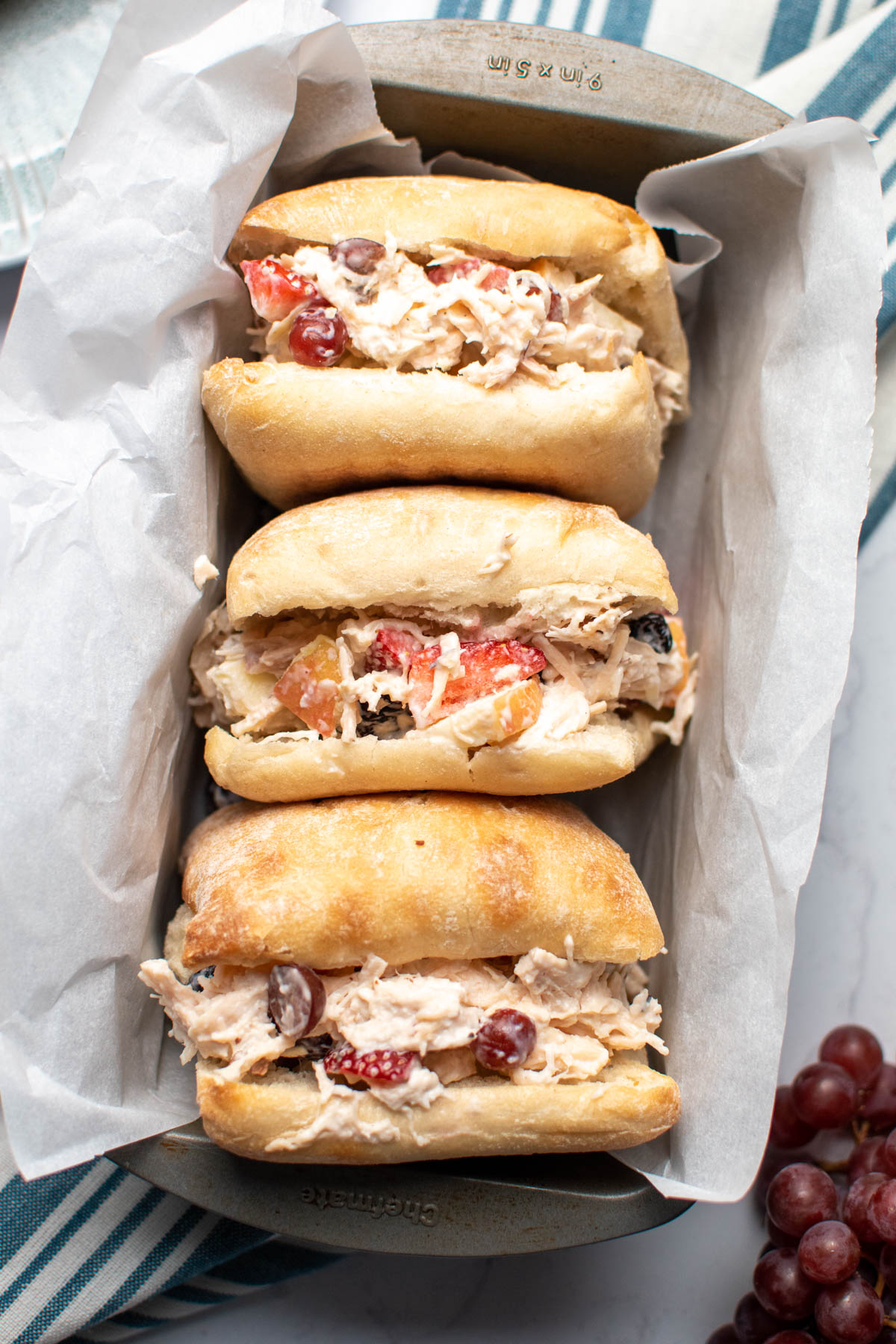 Three chicken salad sandwiches with grapes and apples in parchment paper lined pan.