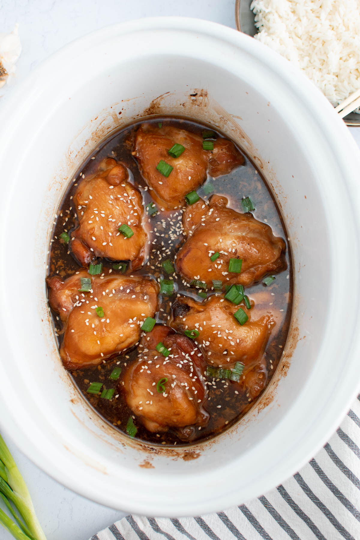 Asian chicken thighs in white Crock Pot on white table with other ingredients nearby.