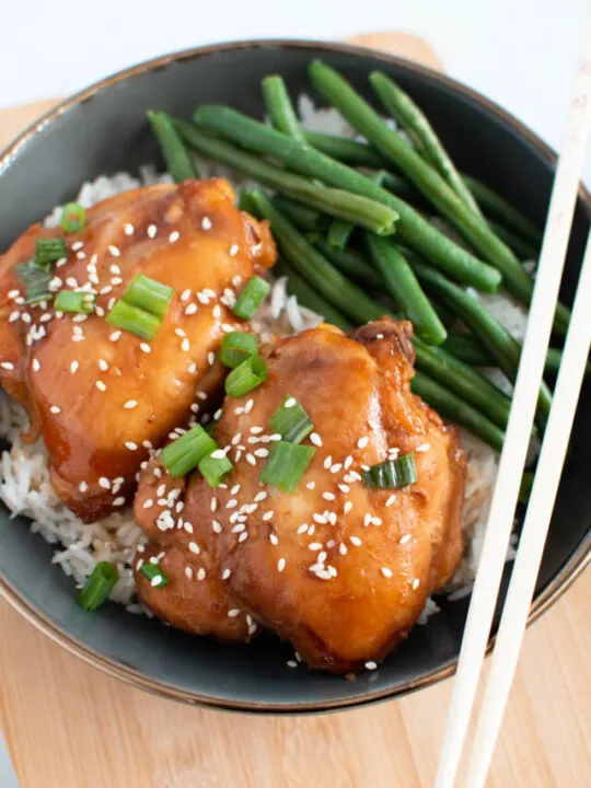 Asian chicken thighs garnished with green onion and sesame seeds in bowl with green beans and rice.