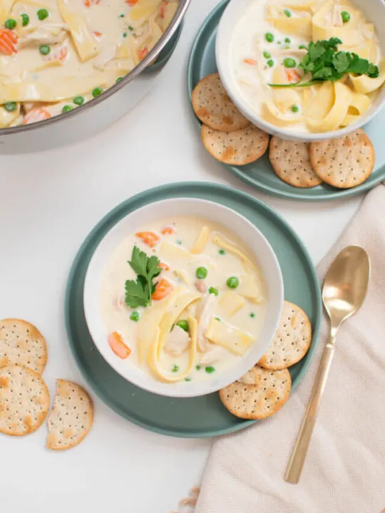 Two bowls of creamy chicken noodle soup on green plates with crackers and spoon nearby.
