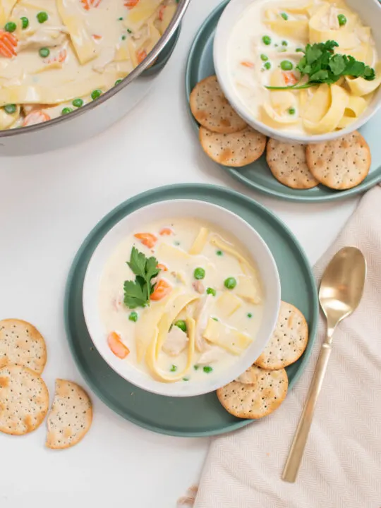 Two bowls of creamy chicken noodle soup on green plates with crackers and spoon nearby.