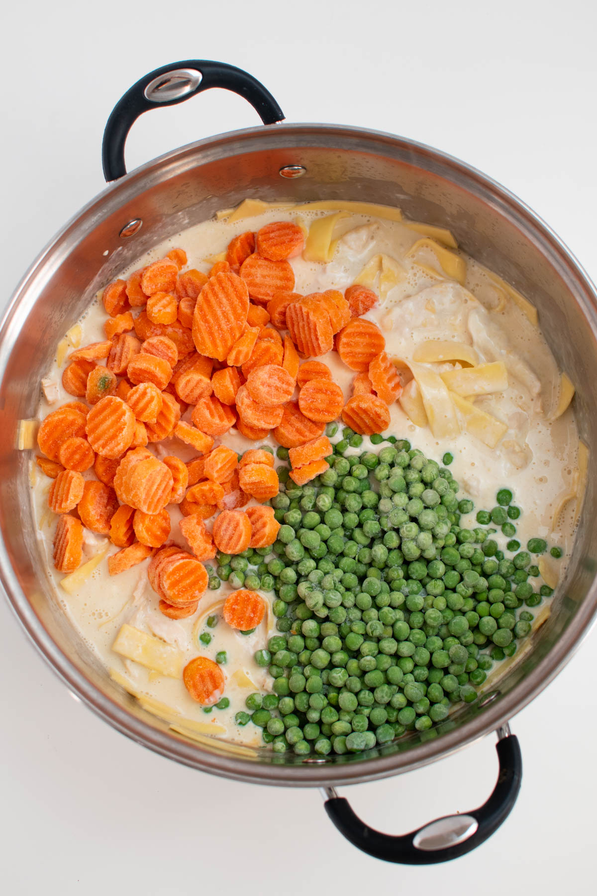 Frozen carrots and peas on top of creamy chicken noodle soup in large pot.
