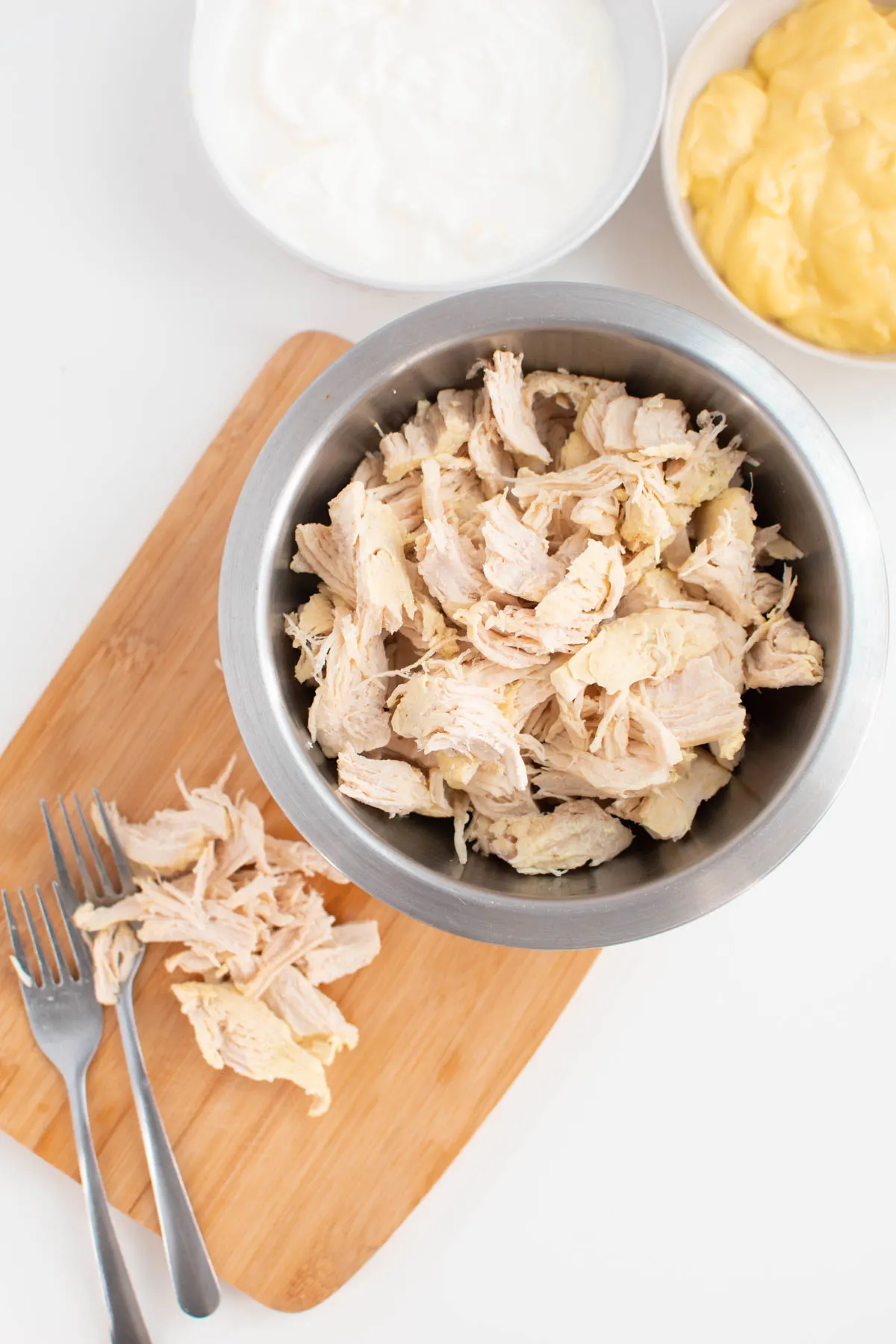 Shredded chicken in small bowl on top of wood cutting board next to bowls of ingredients.