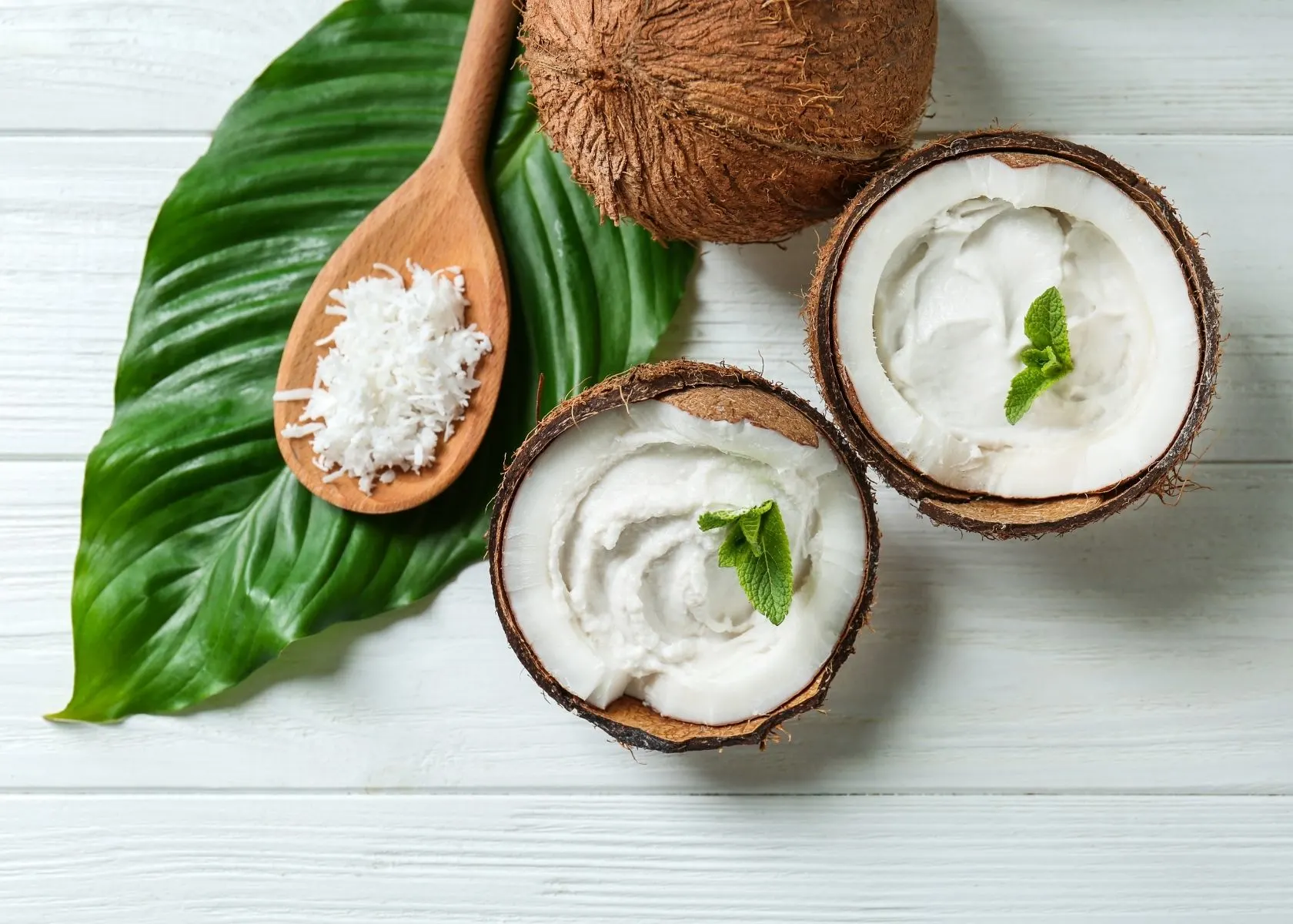 Coconut cream in coconut shells next to large green leaf and shaved coconut.