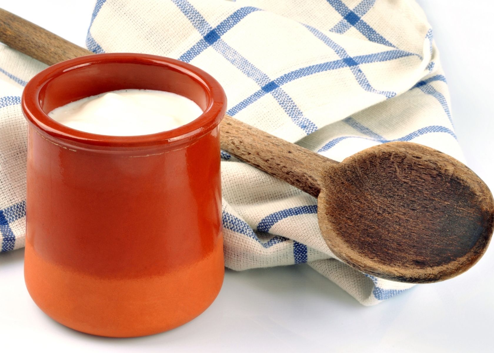 Creme Fraiche in clay mug next to wooden spoon and kitchen towel.