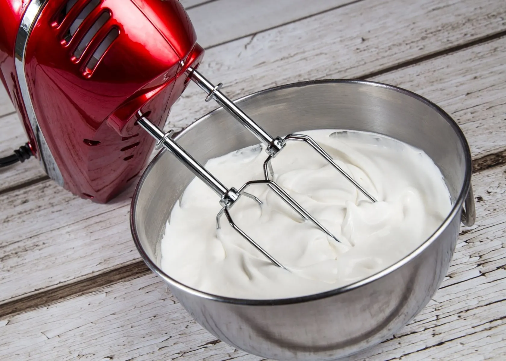 Heavy whipping cream in large metal mixing bowl with red hand mixer.