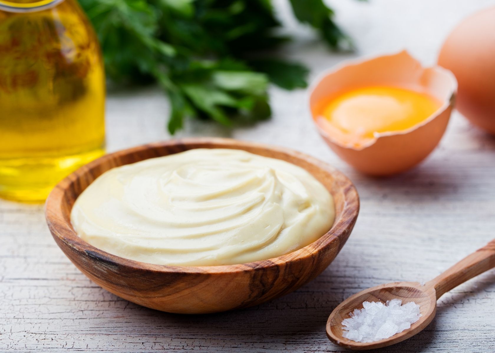 Homemade mayonnaise in wooden bowl next to recipe ingredients like egg, oil and salt.