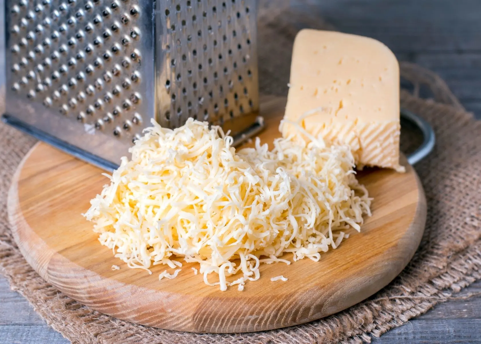 Grated cheese on circular wooden cutting board next to block and metal grater.