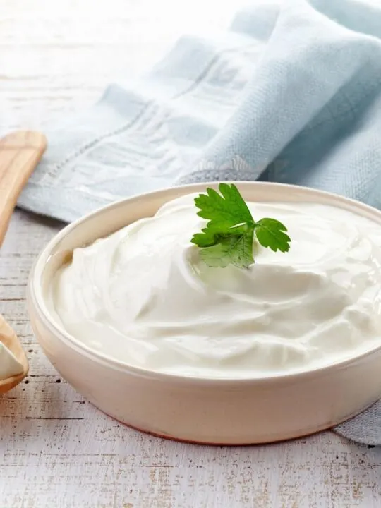 Sour cream in bowl with green garnish next to wooden spoon and kitchen towel.