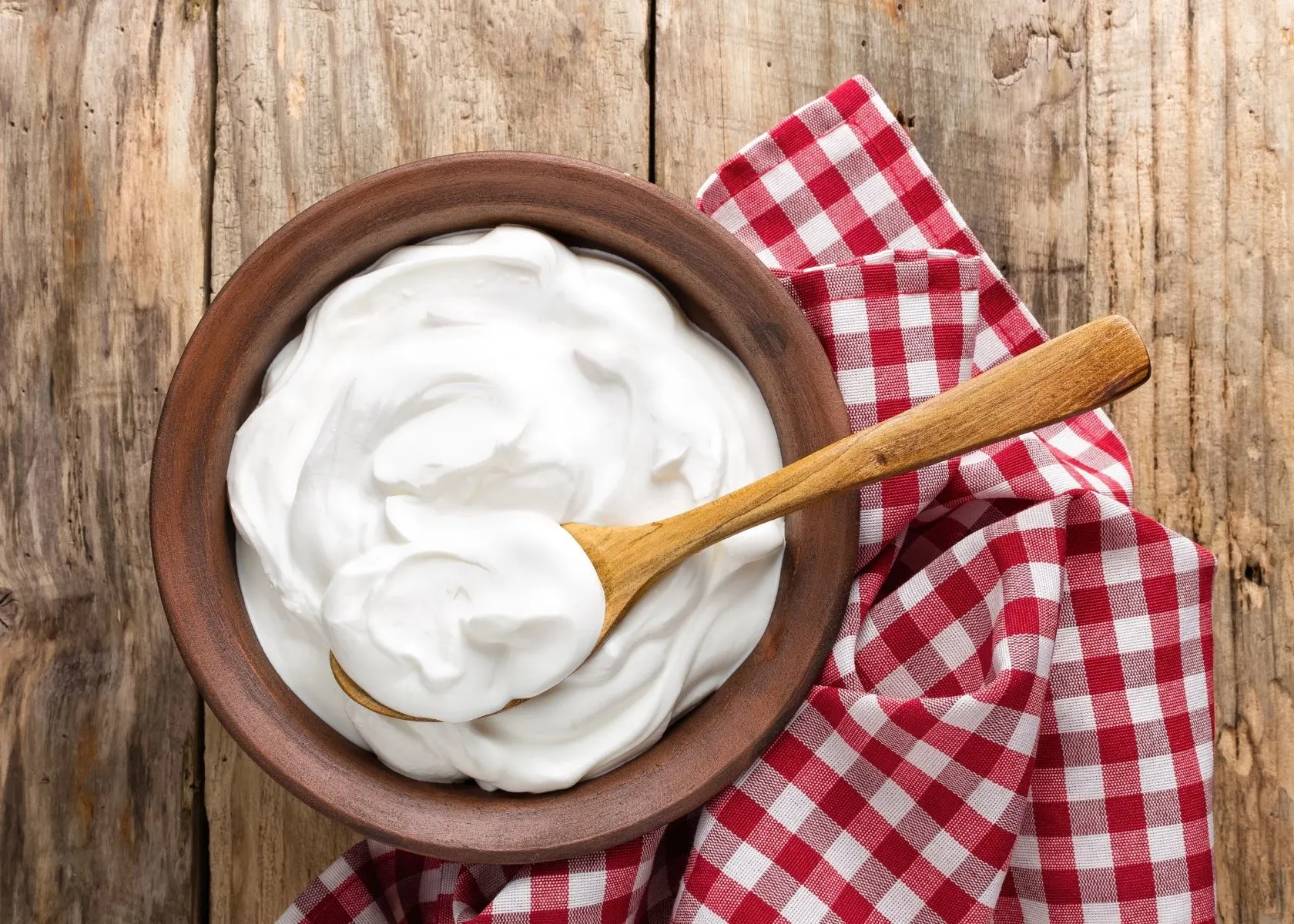 Sour cream in wooden bowl with red plaid kitchen towel and large wooden spoon.
