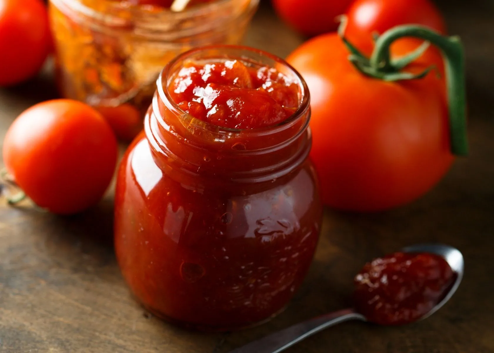 Tomato jam in small clear glass jar surrounded by vine ripened tomatoes.