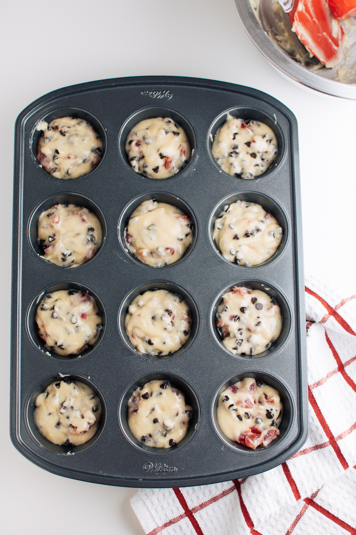 Cherry chip muffin batter in muffin tin next to red and white kitchen towel.
