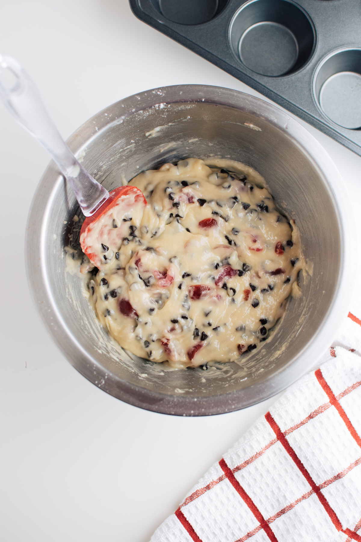 Cherry chocolate chip muffin batter in metal mixing bowl with red and clear plastic spatula.
