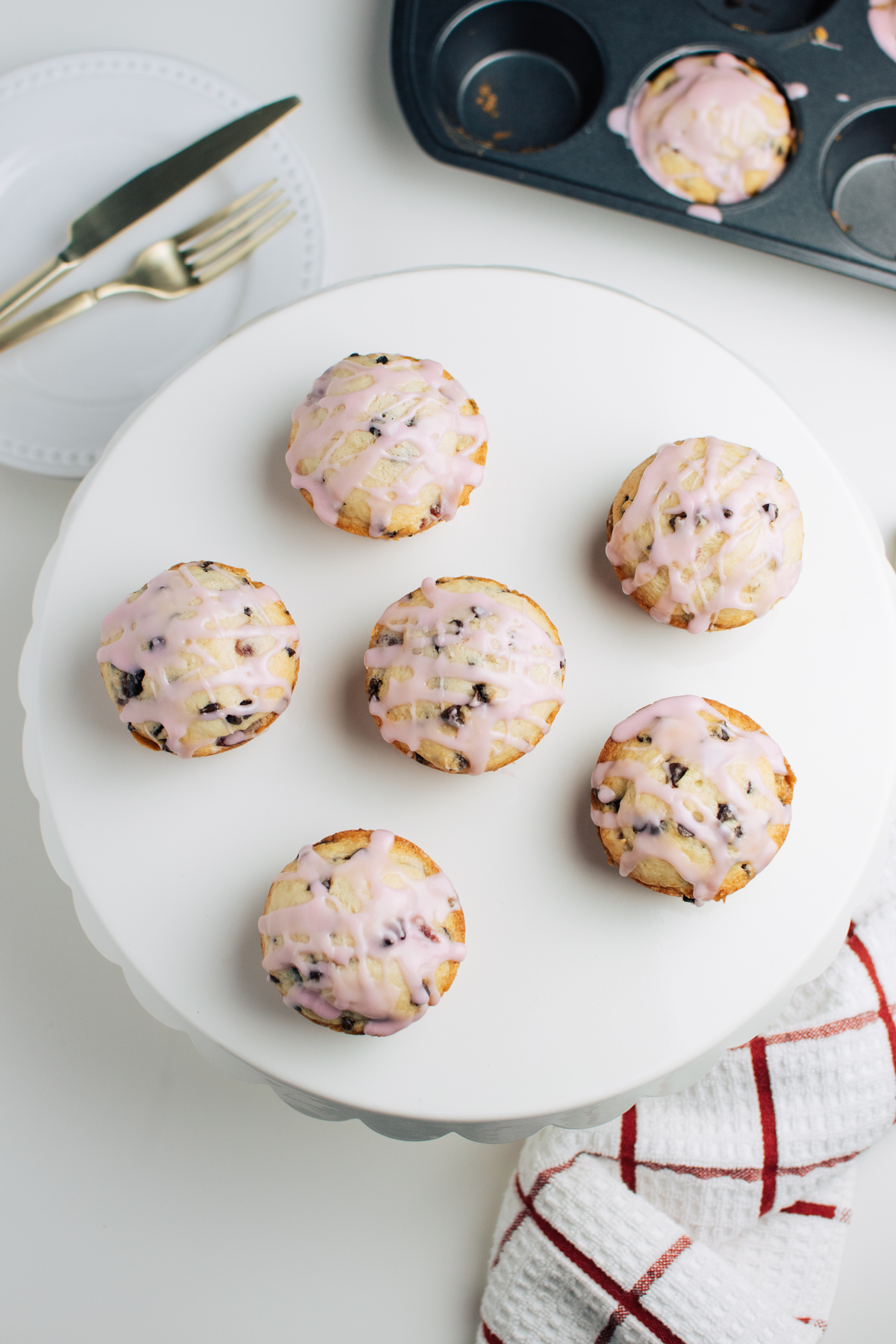 Cherry muffins with pink glaze on large white cake stand next to muffin tin.