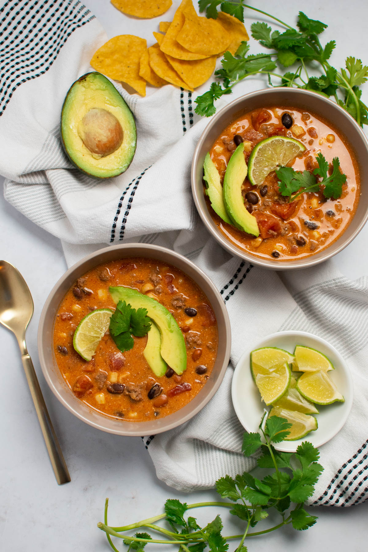 Two bowls of cream cheese taco soup with avocado, lime, and cilantro garnish on top and nearby the bowls.