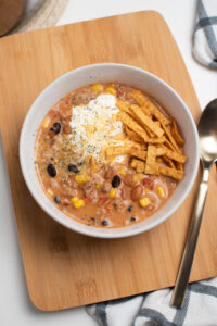 Creamy taco soup in white bowl with tortilla strips and sour cream on wooden cutting board.