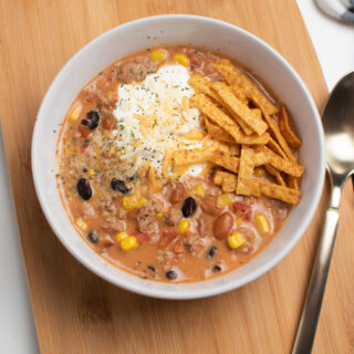 Creamy taco soup in white bowl with tortilla strips and sour cream on wooden cutting board.