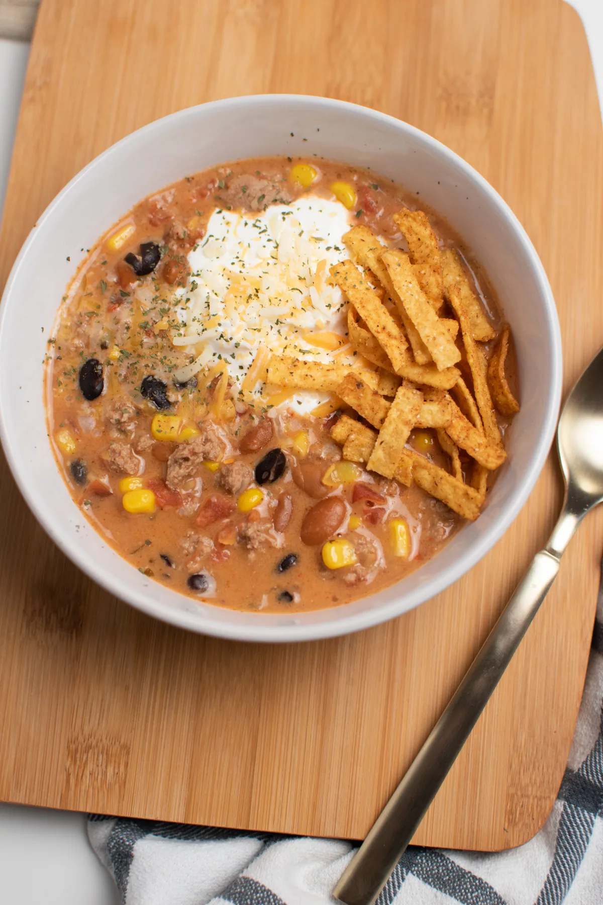 Bowl of taco soup topped with tortilla strips and sour cream on wooden cutting board.