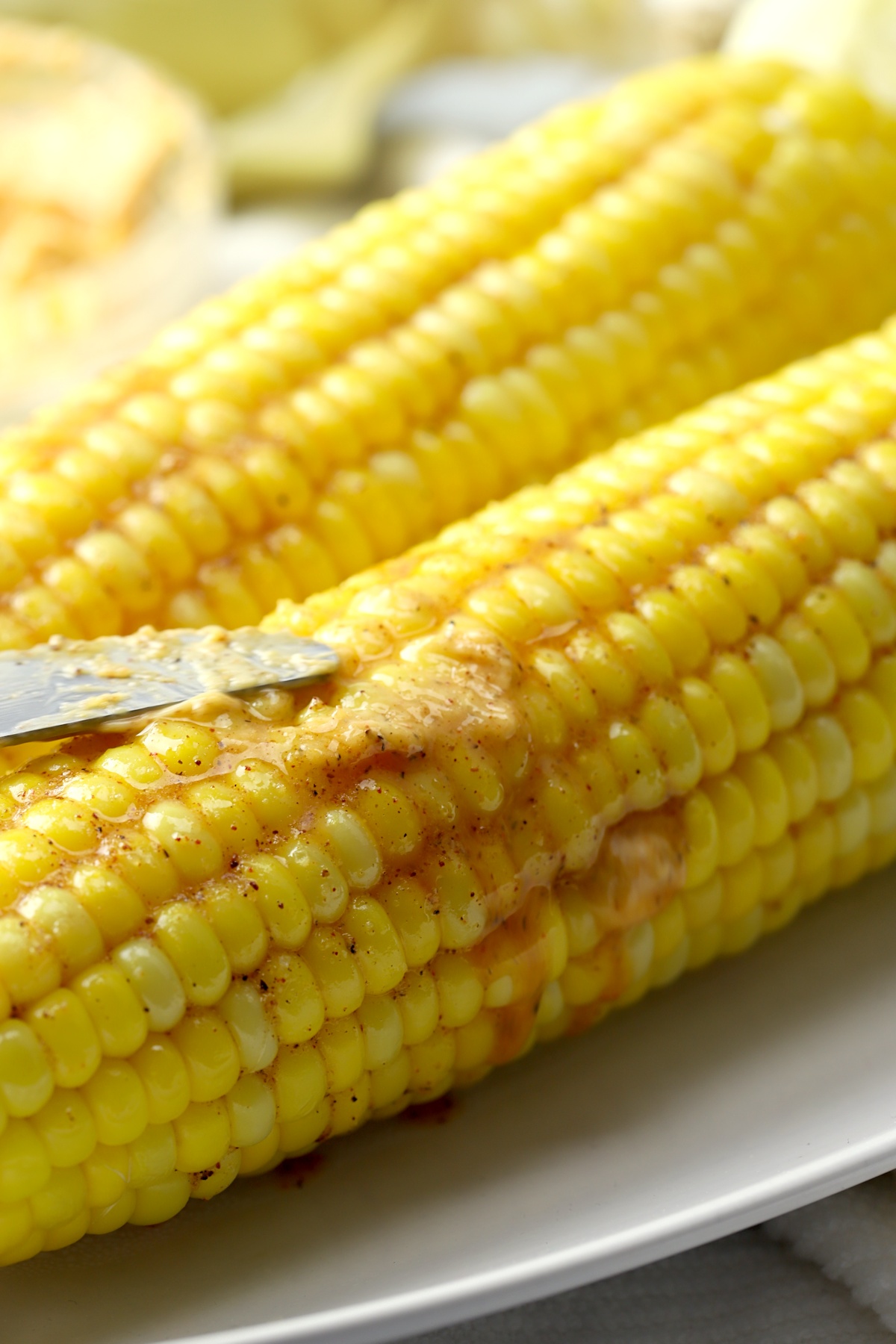 Knife spreads melted butter over oven roasted corn on the cob on a plate.