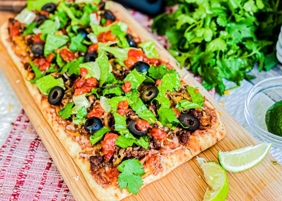 Taco pizza on a cutting board topped with olives and cilantro leaves.