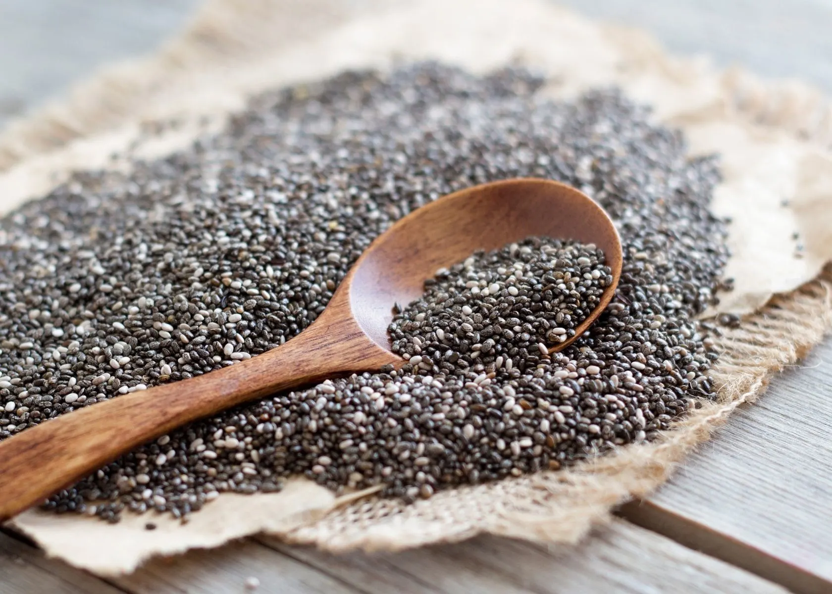 Large pile of chia seeds on burlap cloth with large wooden spoon.