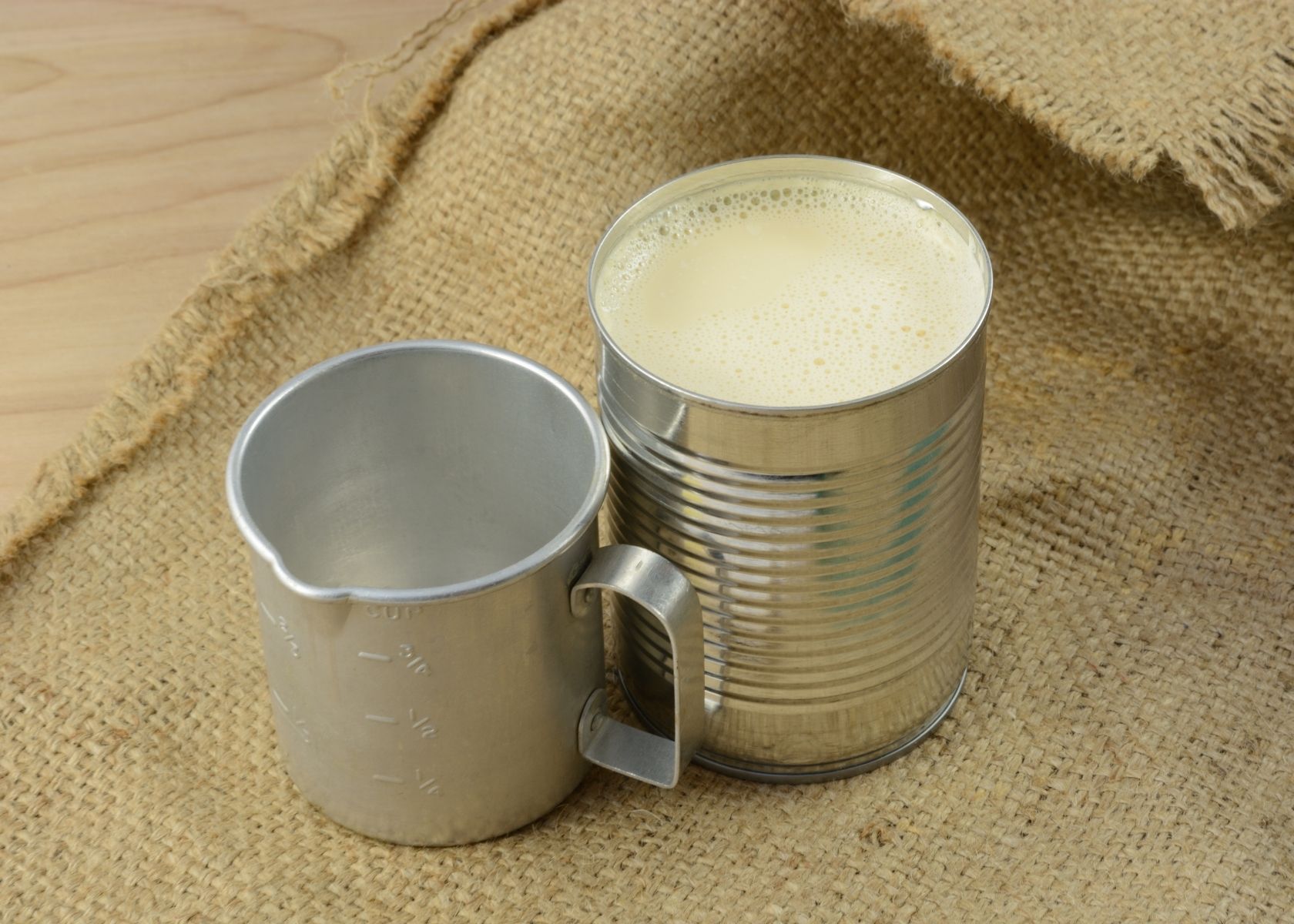 Evaporated milk in can next to measuring cup on a burlap cloth.