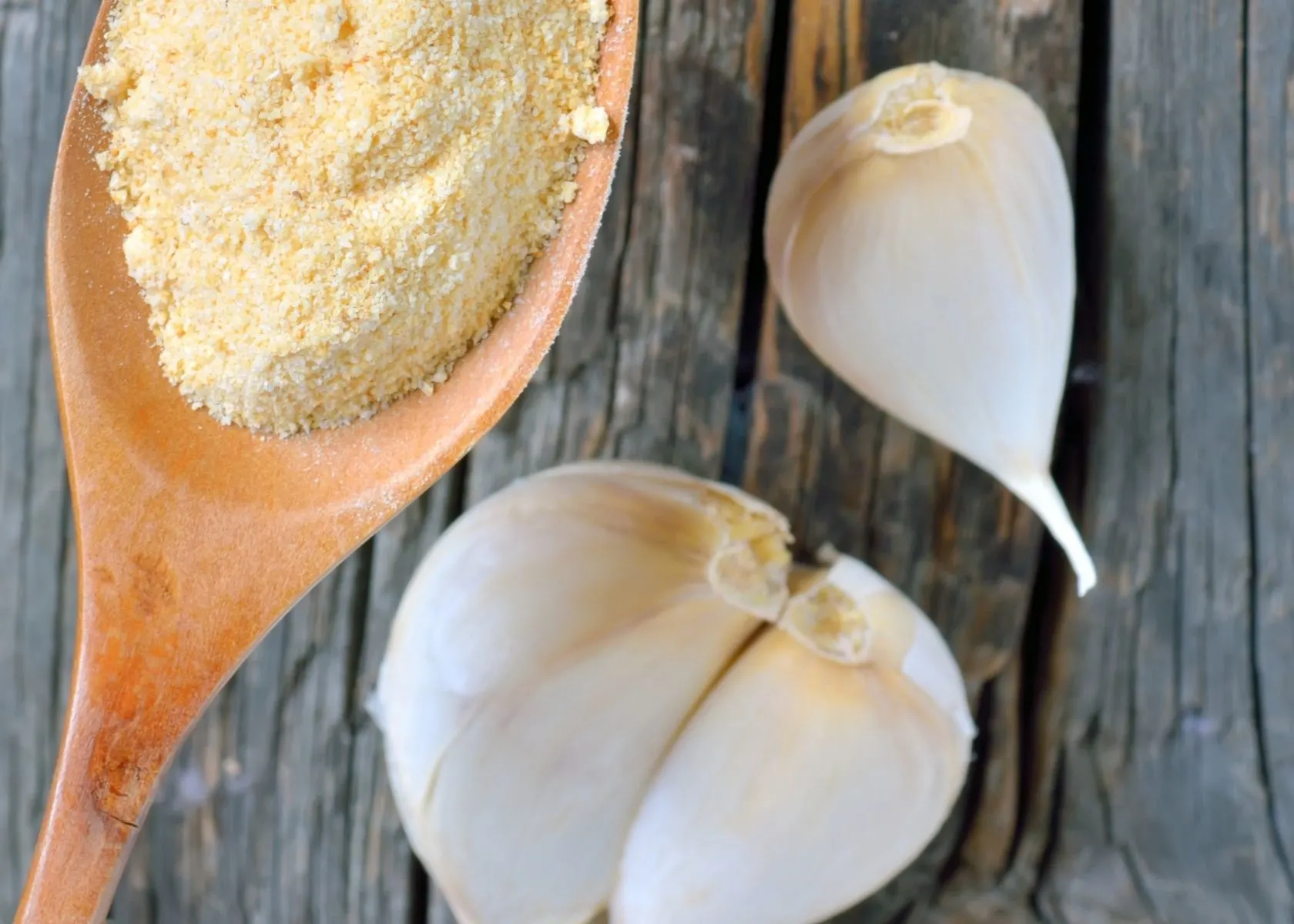 Garlic powder on large wooden spoon next to three fresh garlic cloves.