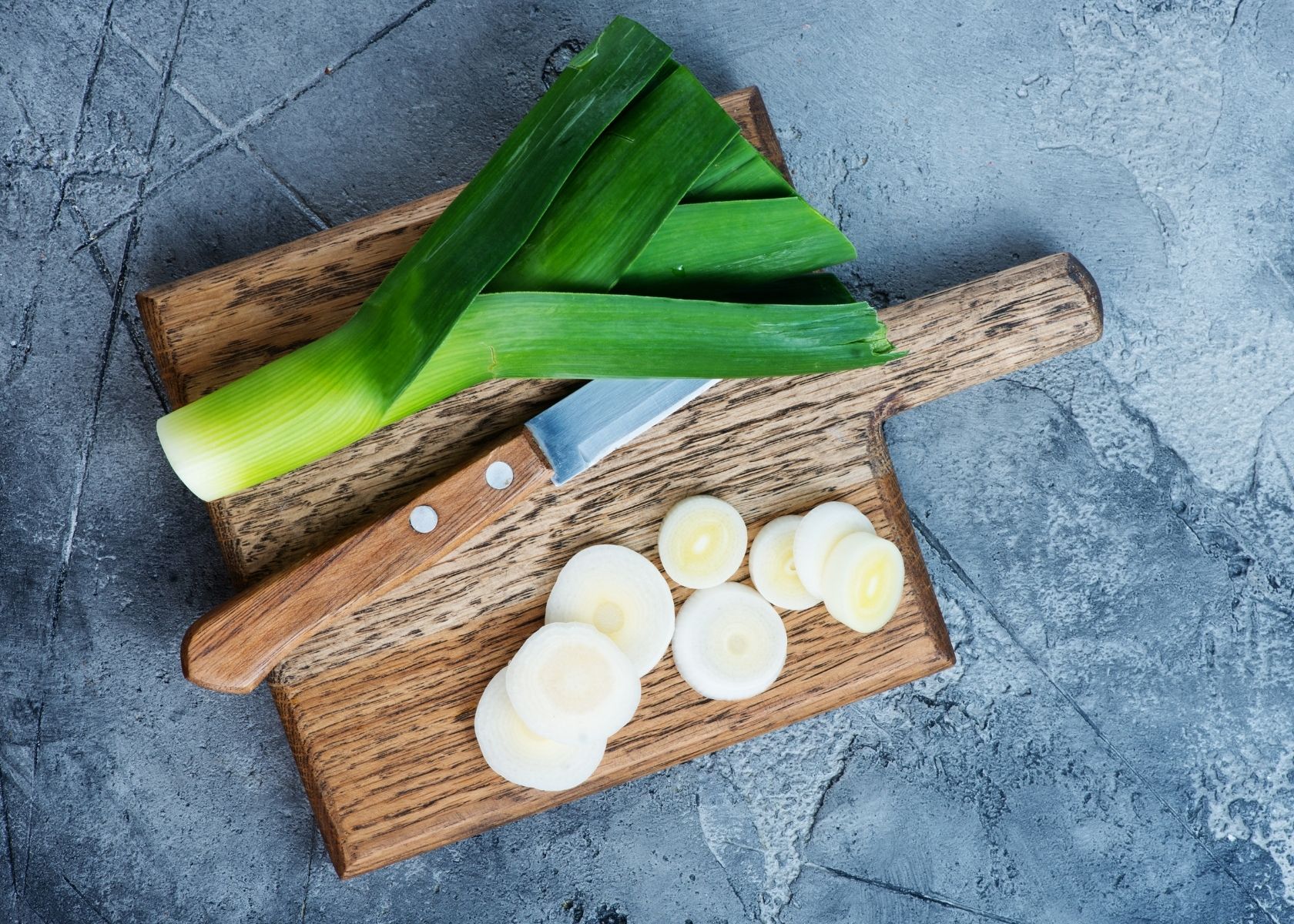 Fresh leek stalk next to cut leek slices on wooden cutting board.