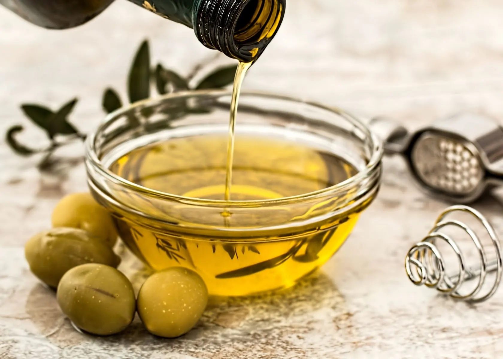 Olive oil is poured into small glass kitchen prep bowl next to fresh olives.