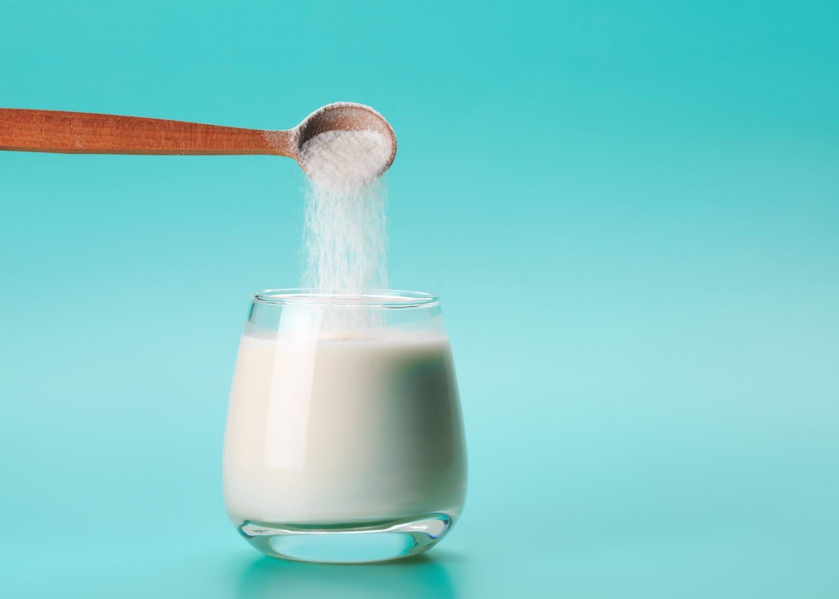 Powdered milk is poured into glass of liquid milk with tiny wooden spoon.