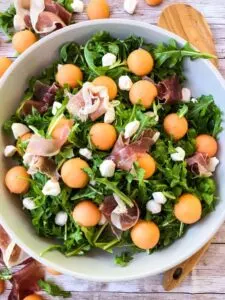 Large white serving bowl filled with cantaloupe prosciutto arugula salad with wooden spoon.