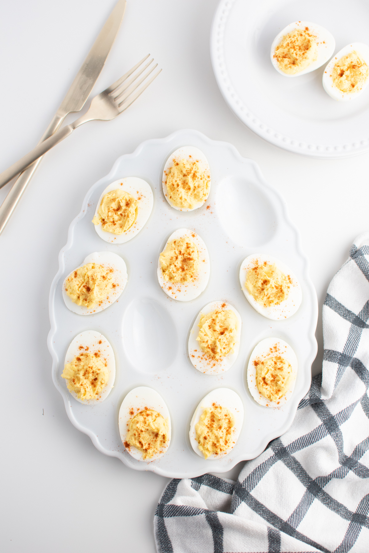 Deviled eggs on white decorative serving platter next to plate of eggs.
