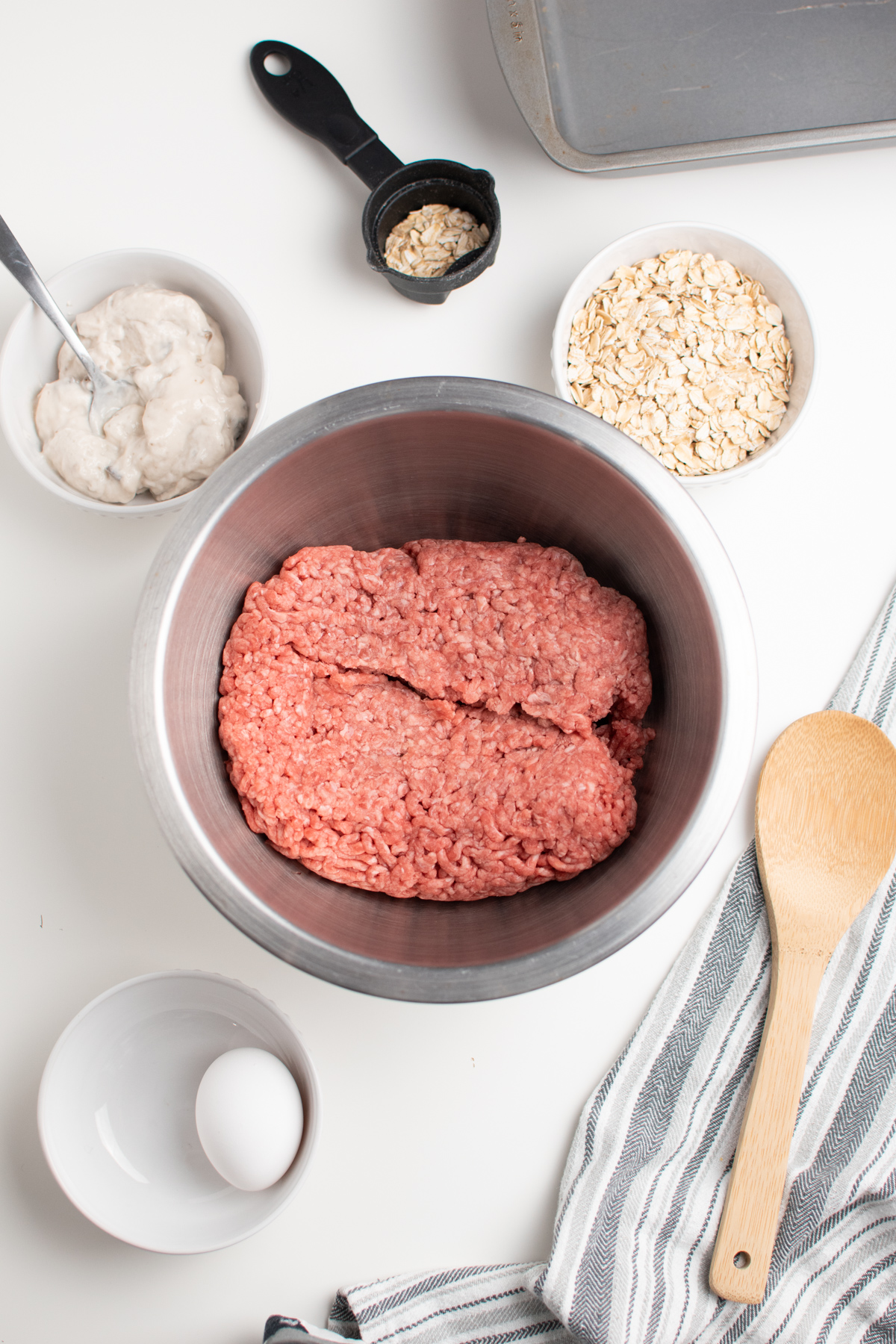 Meatloaf ingredients including ground beef, oats and an egg on white table.
