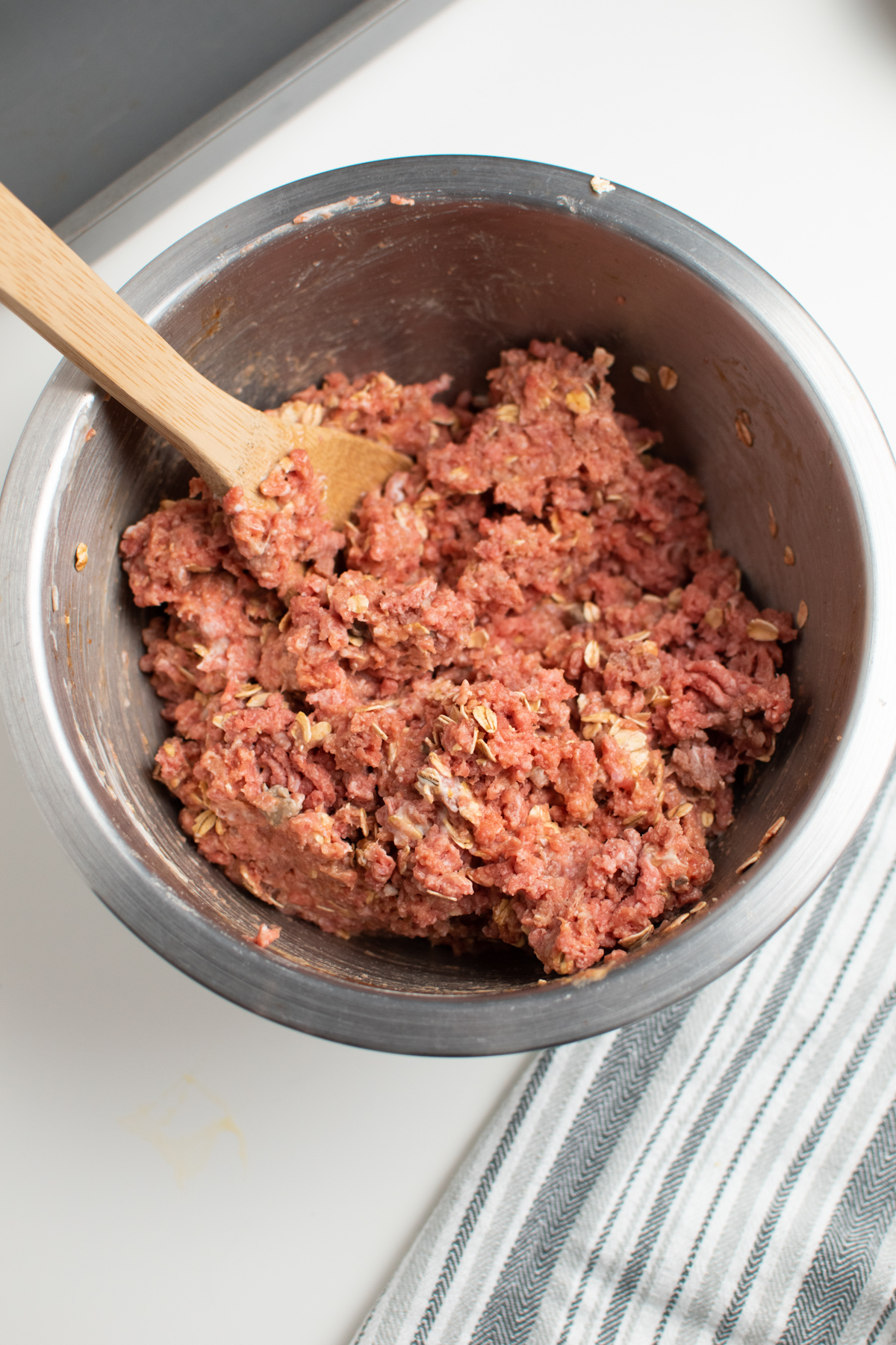 Meatloaf mixture with oats and wooden spoon in large metal mixing bowl.