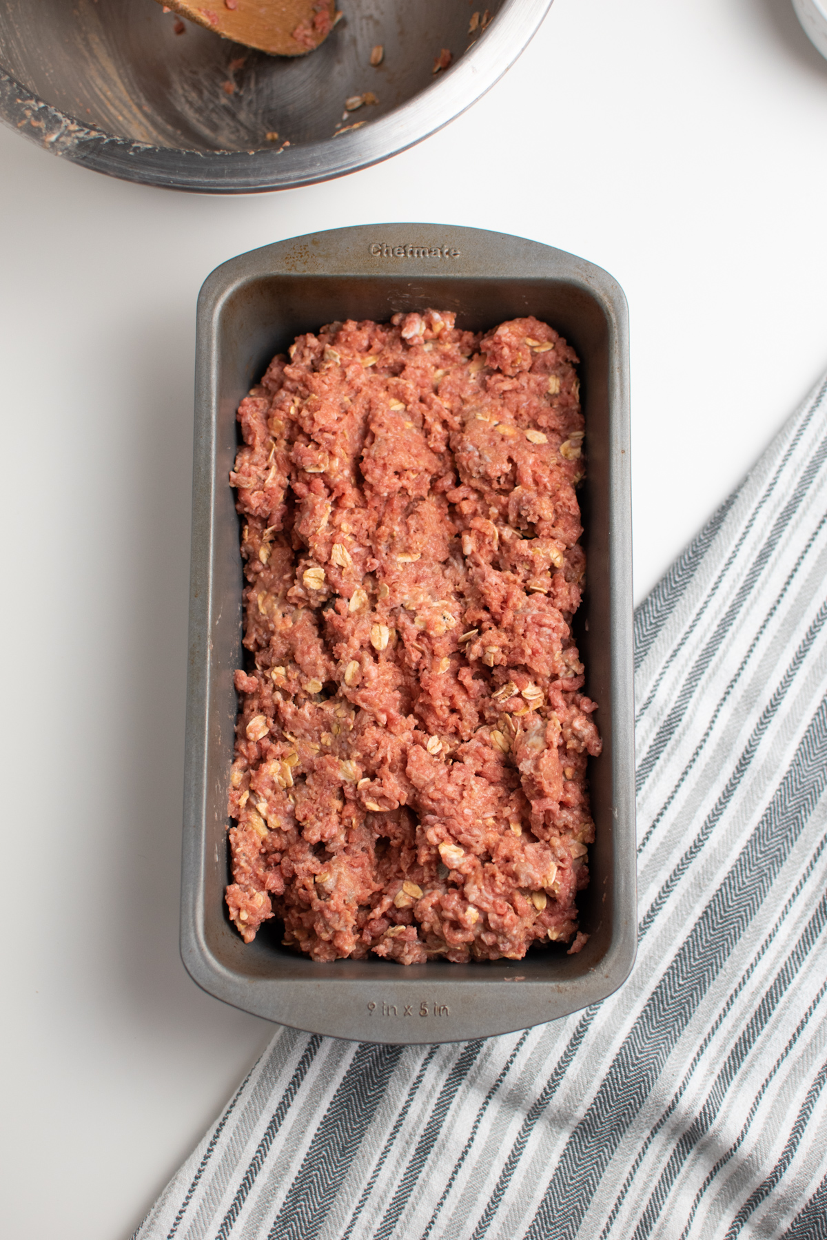 Uncooked ground beef meatloaf mixture in loaf baking pan with kitchen towel.