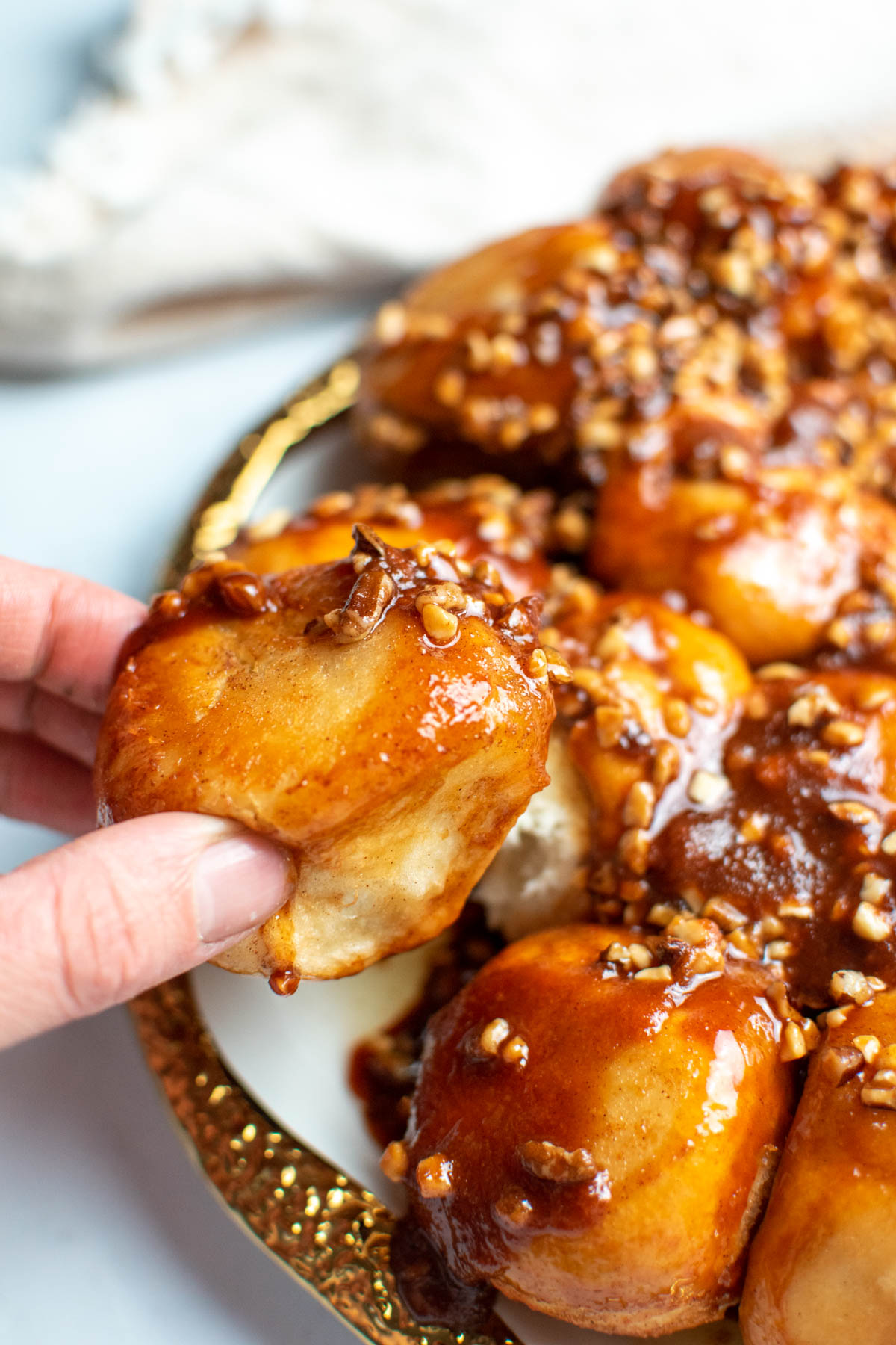 Hand pulls sticky bun with pecans away from other sticky buns on gold plate.