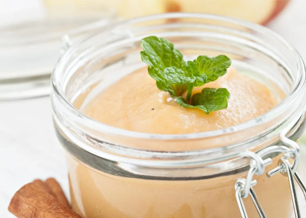 Applesauce in glass jar with mint garnish next to stacked cinnamon pods.