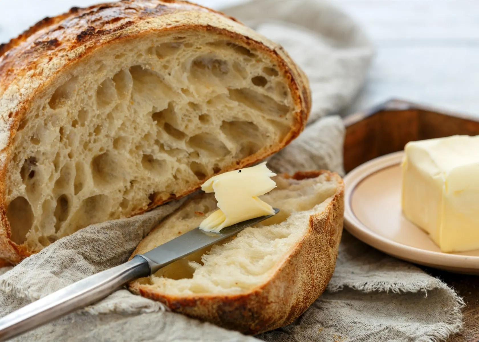 Butter is spread on a loaf of rustic crusty bread next to dish with butter.