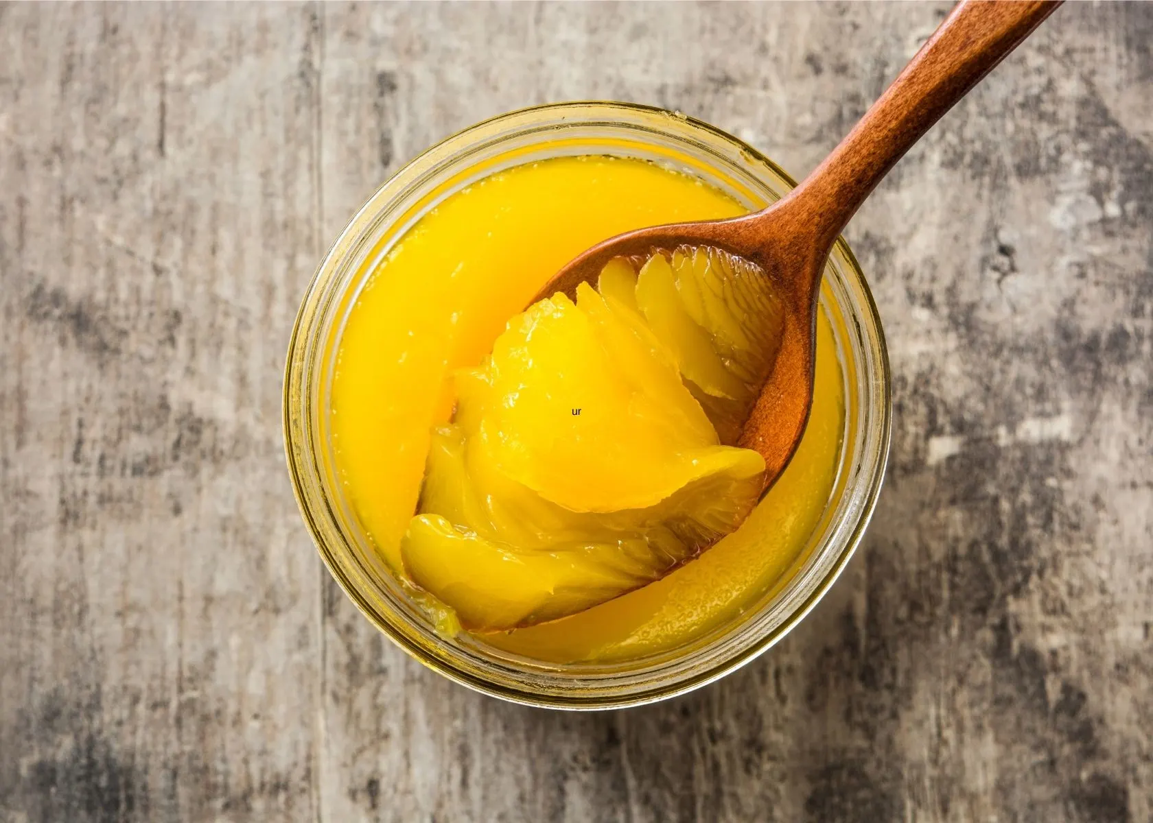 Ghee in clear glass jar with wooden spoon on rustic wooden surface.